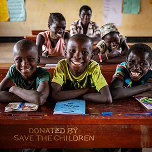 A group of three smiling school boys sit at a desk donated by Save the Children within the Kyangwali Refugee Settlement in Uganda.]